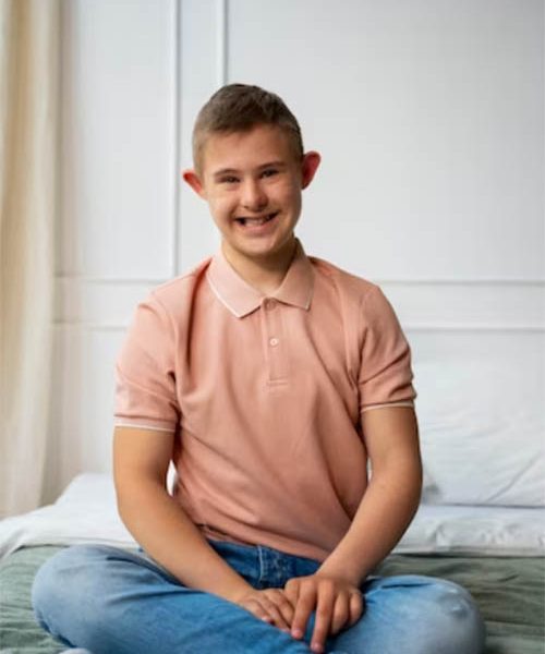 A young man in a pink shirt sitting on a bed.
